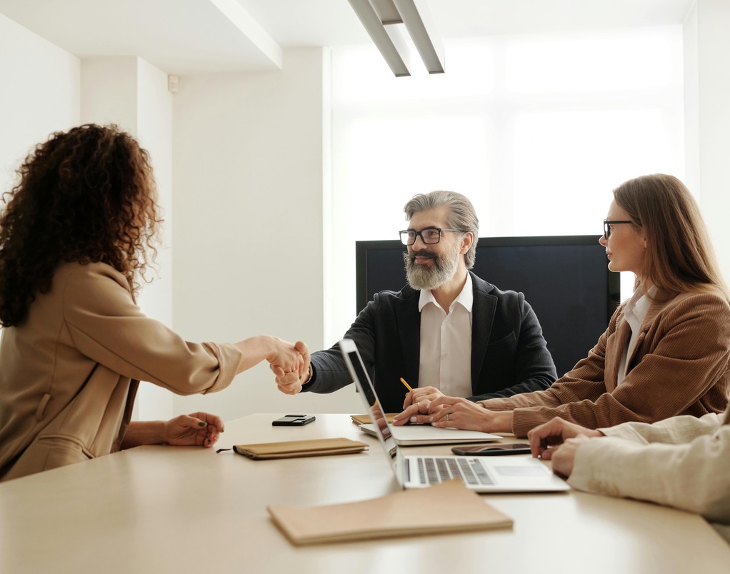 Three professionals in a modern office setting conclude a meeting, with a woman and man shaking hands across a conference table. The scene suggests a successful job interview or networking moment, relevant to career paths in Master of Legal Studies programs. Two other colleagues observe with laptops open, reinforcing a collaborative, business-oriented environment.